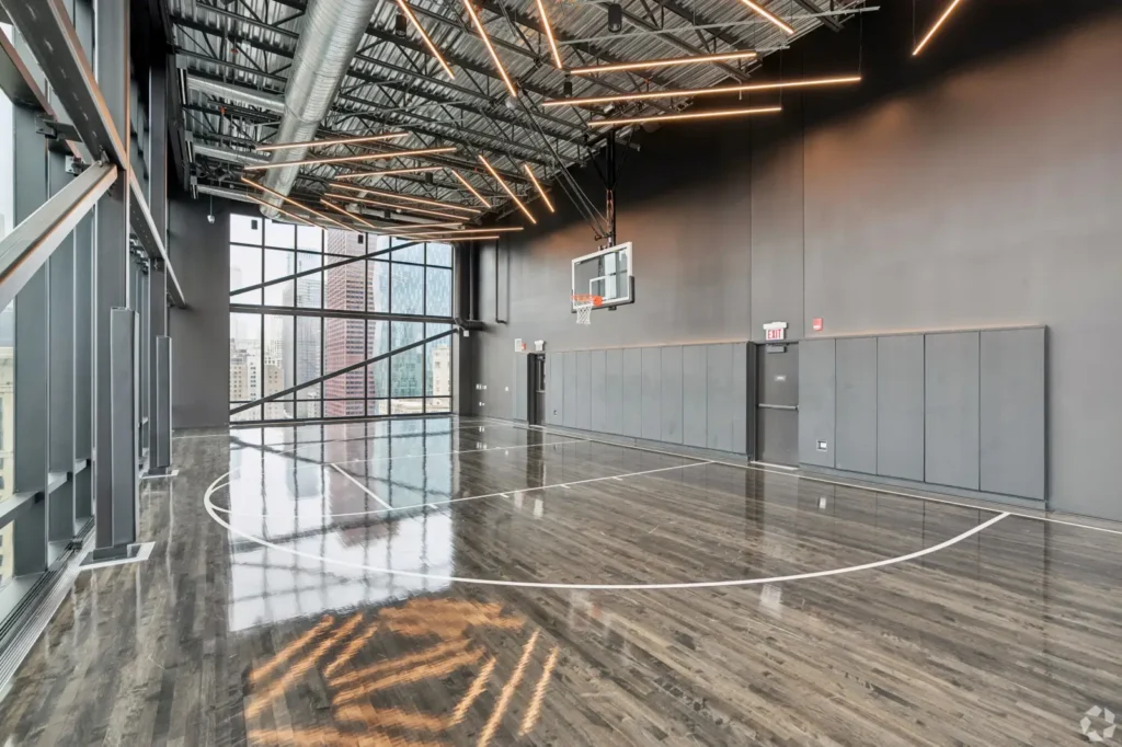 Full indoor basketball court with wood flooring, two hoops, lounge seating, and floor-to-ceiling windows.