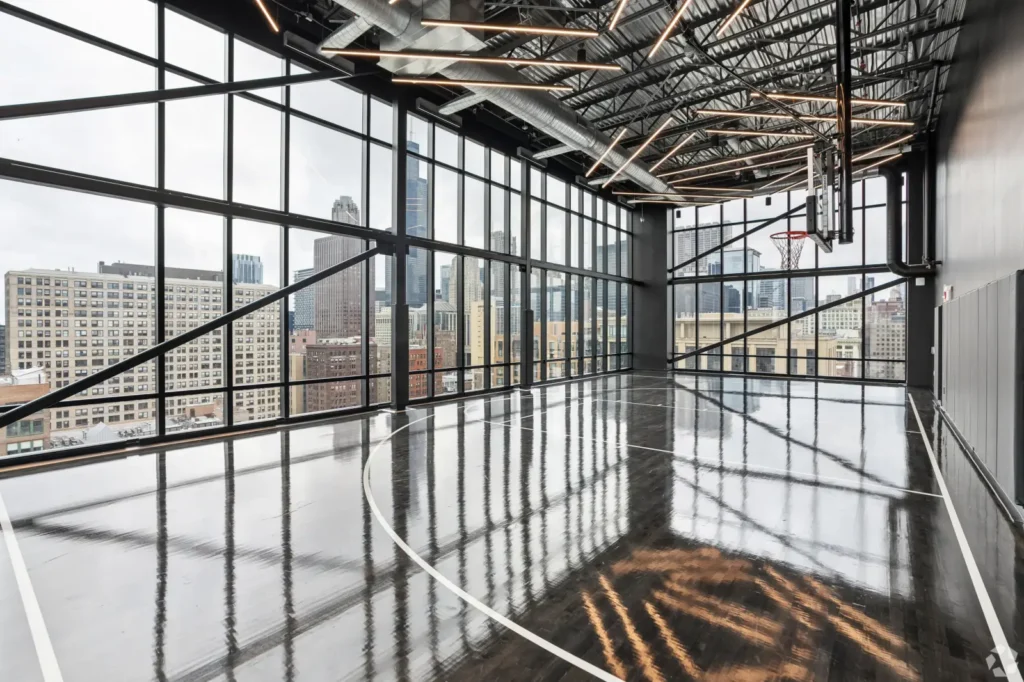 Side view of an indoor basketball court with wood flooring, two hoops, lounge seating, and large windows.