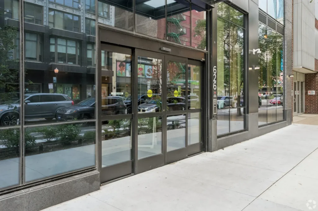 Glass-fronted entrance with revolving doors, landscaped planters, stone walls, and an overhead canopy.