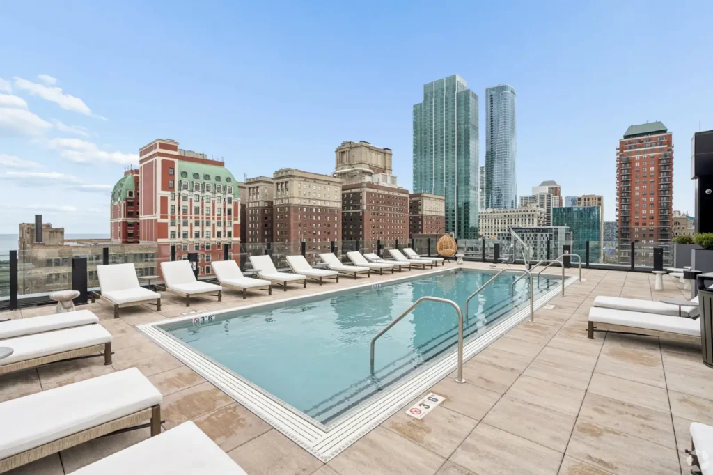 outdoor pool with cushioned lounge chairs, tiled columns, pendant lights, and skyline views.