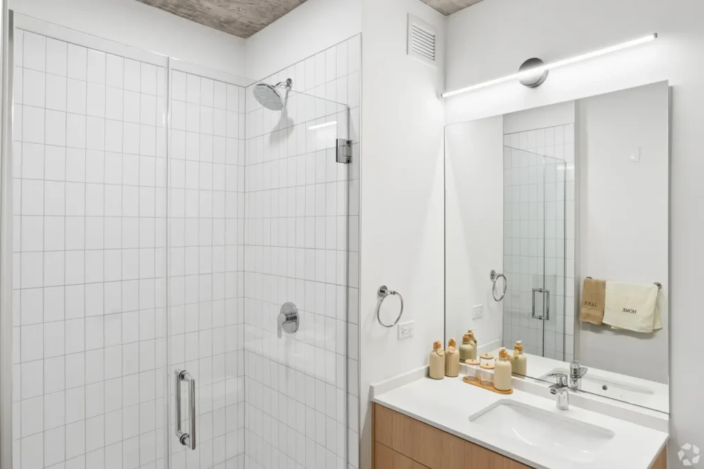 Studio bathroom with a wood vanity, round mirror, glass-enclosed shower, and marble-look tile.