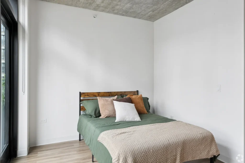 Studio sleeping area with an upholstered bed, nightstand, wall-mounted TV, and floor-to-ceiling windows.